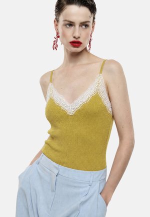 Donna che indossa una canotta in maglia giallo senape con bordo in pizzo bianco e pantaloni azzurri a vita alta, in posa con le mani nelle tasche.