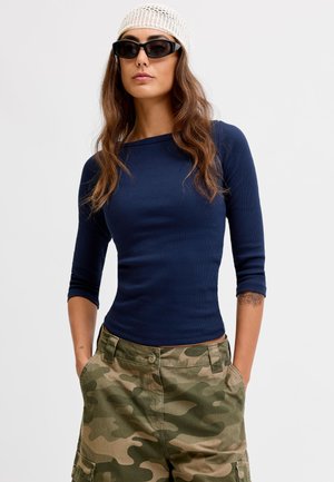 Femme portant un haut bleu marine ajusté, un pantalon cargo camouflage, des lunettes de soleil noires et un bonnet blanc en tricot, les mains dans les poches.