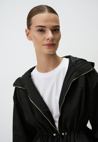 Zwarte hooded jacket met gouden rits en aantrekkoord in de taille, gedragen over een wit t-shirt. Gladde textuur, casual ontwerp.