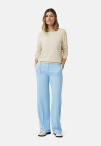 Beiger Langarmpullover mit einer Brusttasche, kombiniert mit hellblauen weit geschnittenen Hosen. Die Schuhe sind in einer Cremefarbe mit schwarzen Sohlen.