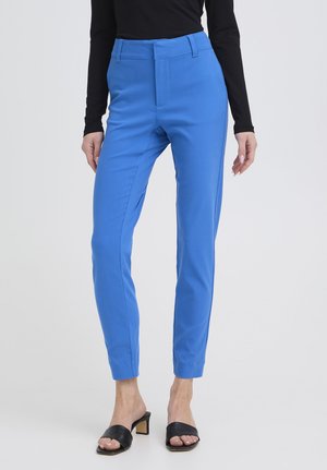 Nainen pukeutuneena kapeamalmaisiin kirkkaan sinisiin nilkkapitkiin housuihin, mustaan pitkähihnaiseen paitaan ja mustiin korkokengillisiin sandaaleihin seisomassa valkoista taustaa vasten.