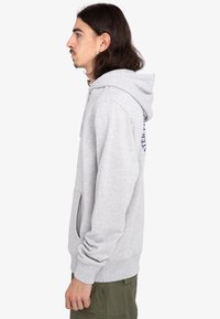 Sudadera gris hecha de tejido suave con capucha ajustable, bolsillo canguro, puños acanalados y aberturas laterales. Presenta texto impreso en la parte posterior.
