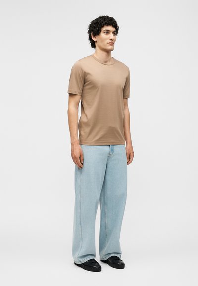 Homme debout portant un t-shirt taupe à manches courtes, un jean large bleu clair et des chaussures noires sur un fond clair uni.