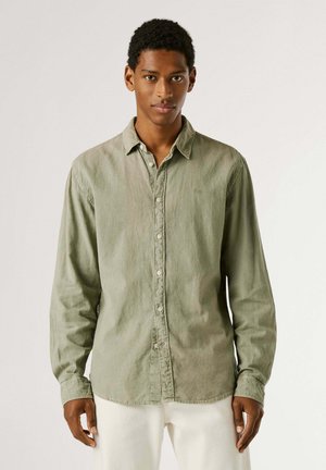 Mladý muž stojaci, oblečený v svetloolivovo zelenej košeli s dlhým rukávom na gombíky a bledých nohaviciach na jednofarebnom pozadí.