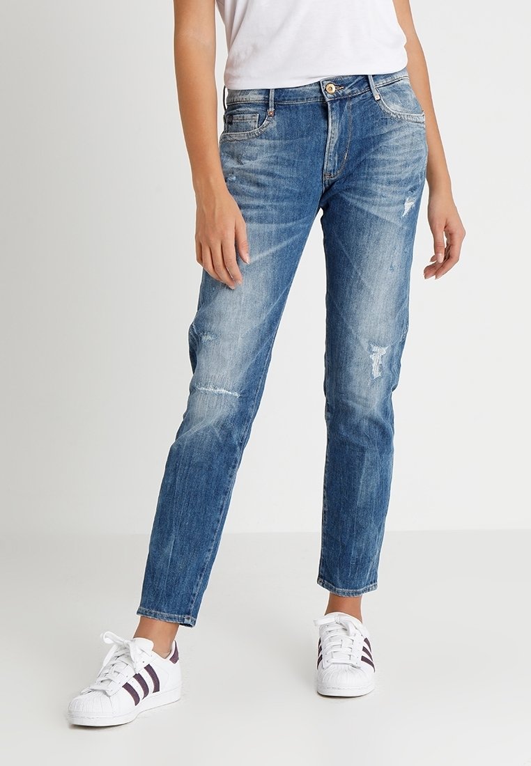 Vaqueros de denim azul claro con corte ajustado, que presentan áreas desgastadas y desvanecidas. Combinados con zapatillas blancas con acentos morados.
