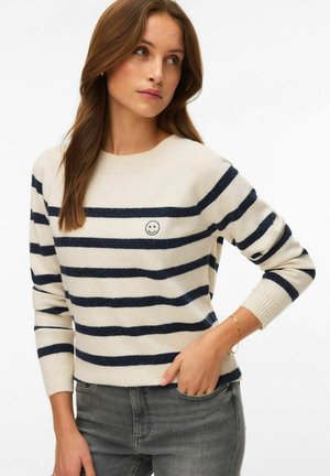 Femme portant un pull crème à rayures horizontales bleu marine avec un petit emblème visage souriant, associée à un jean gris, debout devant un fond blanc.