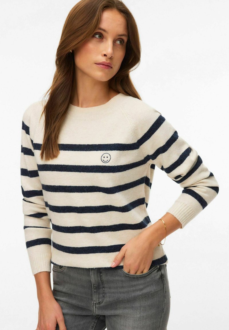 Femme portant un pull crème à rayures horizontales bleu marine avec un petit emblème visage souriant, associée à un jean gris, debout devant un fond blanc.