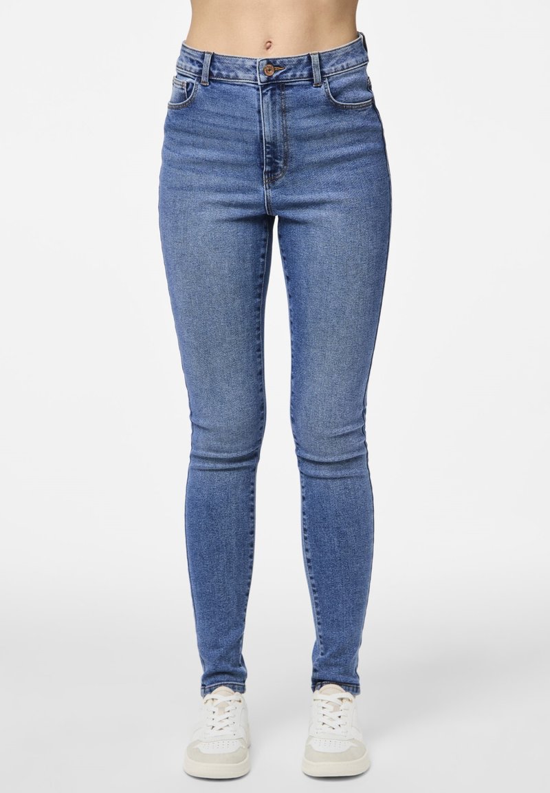 Femme portant un jean skinny bleu taille haute et des baskets blanches, se tenant devant un fond clair.