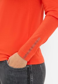Langsleeve oranje shirt met een ronde halslijn en een rij strasstenen op de mouwzoom. Draagt met een grijze spijkerbroek, hand in de zak.