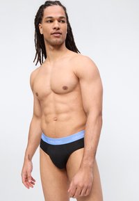 Moški brez srajce z dolgimi kitkami v črnih spodnjicah Calvin Klein z svetlo modrim pasom, ki gleda levo.