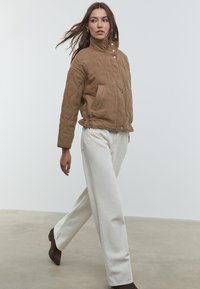 Braune gesteppte Jacke mit hohem Kragen, Druckknöpfen und Seitentaschen, kombiniert mit weißen weiten Hosen und braunen Ankle Boots.