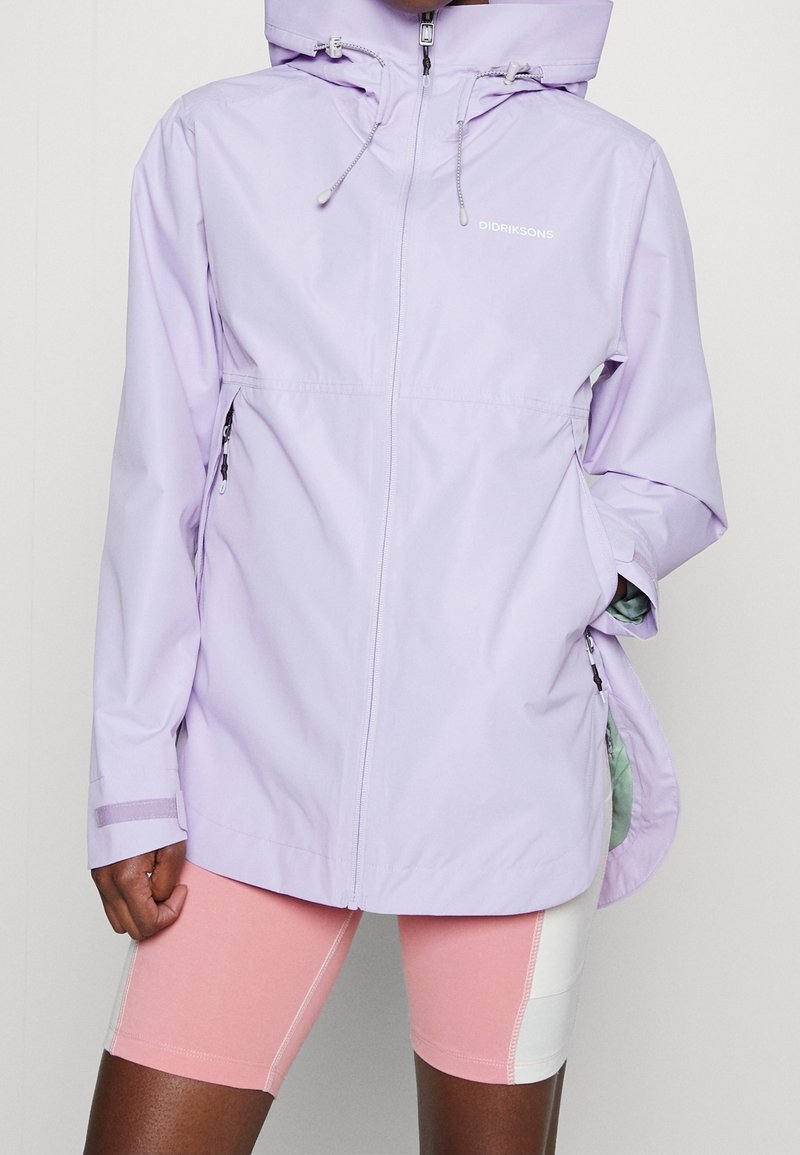 Personne portant une veste à capuche violet clair avec une fermeture éclair et des cordons, associée à un cuissard de cyclisme rose et blanc.