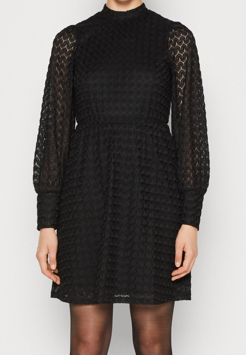 Femme portant une robe longue noire à manches longues en dentelle texturée, avec un col montant et des poignets élastiqués, vue du cou jusqu'à mi-cuisse.