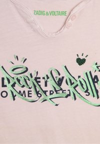 Lyserød T-shirt med rund udskæring, prydet med grøn metallic graffitistil tekst og sort skrift. Inkluderer lille grønt hjerte-detalje.