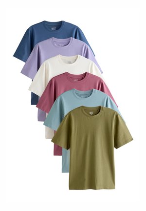 Mand med kort krøllet hår iført en olivengrøn T-shirt med korte ærmer og blå jeans står mod en ensfarvet hvid baggrund.
