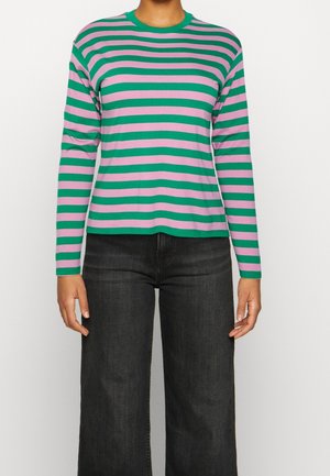 Person iført en langærmet grøn og lilla trøje med horisontale striber kombineret med mørke, højttaljede bredbenede jeans mod en hvid baggrund.