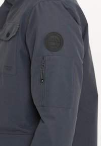 Marineblå jakke med en lynlåslomme på ærmet, brystlomme og et rundt gummilogo-patch på overarmen. Glat, slidstærkt stof.