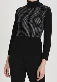 Sweater noir à col roulé avec une partie supérieure texturée et à motifs comportant des points blancs ; design côtelé sur le bas du corps et les manches.