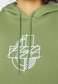 Personne portant un sweat à capuche vert avec des cordons et un design graphique abstrait blanc présentant des formes géométriques et un texte en écriture cursive sur la poitrine.