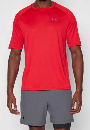 Mann trägt ein hellrotes kurzärmeliges Sportshirt und graue Under Armour-Shorts vor einem schlichten Hintergrund.