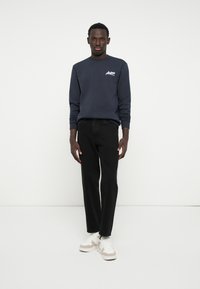 Marine crewneck sweatshirt, zwarte jeans en witte sneakers met beige accenten. Model staat tegen een egale witte achtergrond, handen aan de zijkanten.