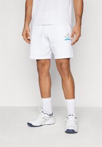 Hvide atletiske shorts med Lotto-logo i blå og gul, parret med hvide sokker og sneakers med marineblå accenter. Glat stofstruktur.