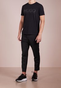 Mann trägt ein schwarzes Hugo Boss T-Shirt, schwarze kurze Hosen und schwarz-weiße Sneakers und steht vor einem einfarbigen beigen Hintergrund.