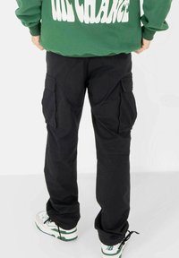 Zwarte cargobroek met meerdere zakken, een losse pasvorm en een gladde stoftextuur. Draagt een groene sweatshirt en witte sneakers.
