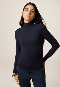 Navy coltrui van zachte brei stof, met knoopaccenten op de schouder en een aangesloten ontwerp. Gecombineerd met donkere broeken.