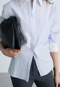 Camicia button-up a righe blu chiaro con un grande colletto, piccola tasca frontale e maniche arrotolate, abbinata a una pochette in pelle nera.