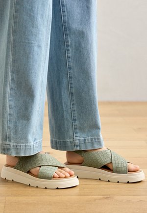 Green, crossband sandals with textured surface, light cushioning, and white platform sole. Paired with light blue wide-leg jeans on wooden floor.