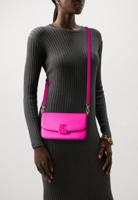 Borsa a tracolla in pelle rosa con design a ribalta, dotata di un logo dorato e di una tracolla sottile, abbinata a un vestito grigio a maniche lunghe con righe.