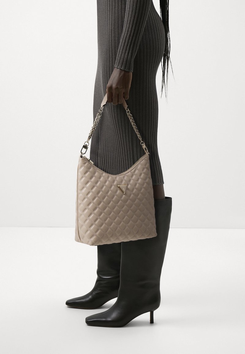 Personne en robe grise côtelée et bottes noires à talons tient un sac à main beige matelassé avec une bandoulière chaîne sur le côté.