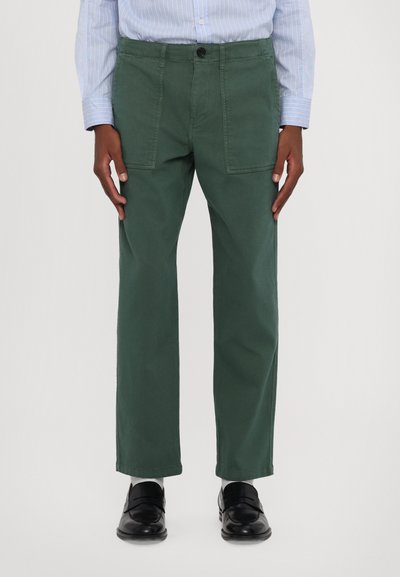 Pantaloni din bumbac verde cu picior drept, având buzunare mari în față, închidere cu nasture și o textură fină. Purtați cu pantofi negri.