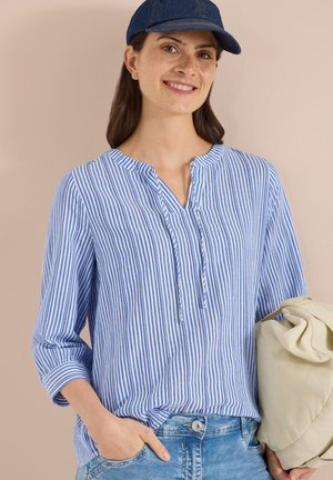 Smilende kvinde iført en blåstribet skjorte, cowboybukser og en navy-kasket, der holder en beige taske mod en ensfarvet beige baggrund.