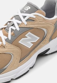 Beige és szürke sportcipő, textúrált hálós felsőrésszel, velúr kiegészítőkkel, fehér fűzőkkel és egy hangsúlyos fehér "N" logóval az oldalon.
