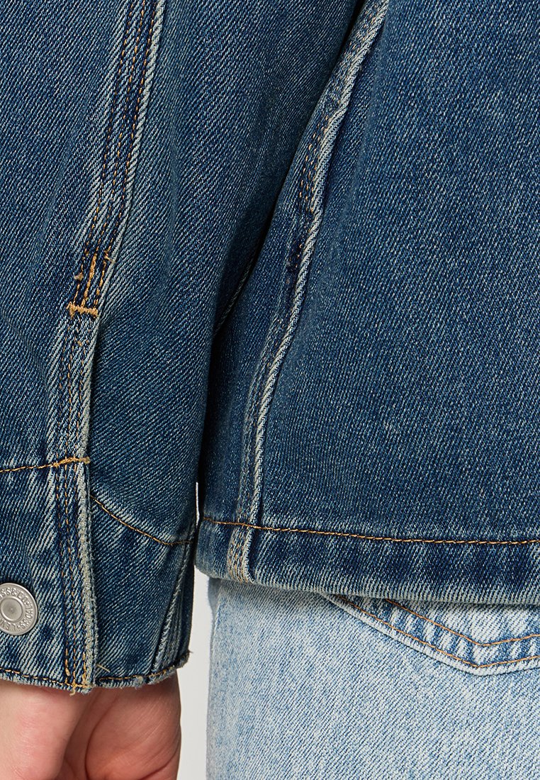 Chaqueta de denim de color azul oscuro, con costuras visibles y un detalle de botón en el puño. La tela tiene una apariencia texturada y tejida.