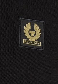 Black fabric with a rectangular patch featuring a gold logo of stylized wings and a circular emblem, labeled "BELSTAFF" at the bottom.