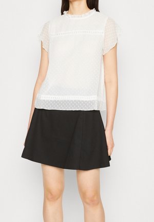 Vrouw die een doorschijnende witte blouse met korte mouwen draagt, met polka dots en een ruchtrand bij de hals, gecombineerd met een zwarte A-lijn rok.