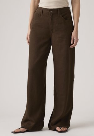 Žena oblečená v hnedých nohaviciach s vysokým pásom a širokými nohavicami a v čiernych žabkách, stojaca pred jednoduchým pozadím.