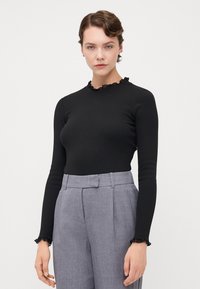 Black long-sleeve top with a textured ruffled neckline, paired with light gray high-waisted trousers featuring sharp creases and side pockets.