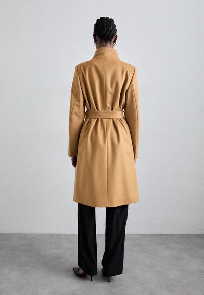 Camel wool-blend coat with a belted waist, long sleeves, and a high collar, paired with black wide-leg trousers and pointed heels.