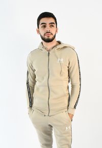 Sudadera beige con cremallera, bolsillo tipo canguro, capucha y rayas laterales negras con texto blanco; combinada con pantalones de chándal a juego.