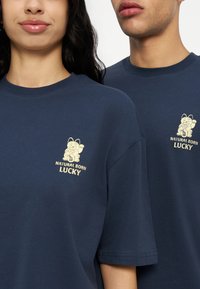 Δύο άτομα που φοράνε μπλουζάκια μπλε ναυτικού με μια μπεζ γατούλα που κυματίζει και κείμενο "ΦΥΣΙΚΑ ΤΥΧΕΡΟΙ" στο στήθος.