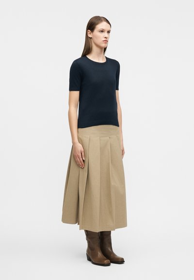 Γυναίκα που στέκεται, φοράει μπλουζάκι κοντομάνικο σε σκούρο μπλε, φούστα midi με πτυχώσεις σε μπεζ χρώμα και καφέ καουμπόικες μπότες με τακούνι, μπροστά σε μονόχρωμο φόντο.