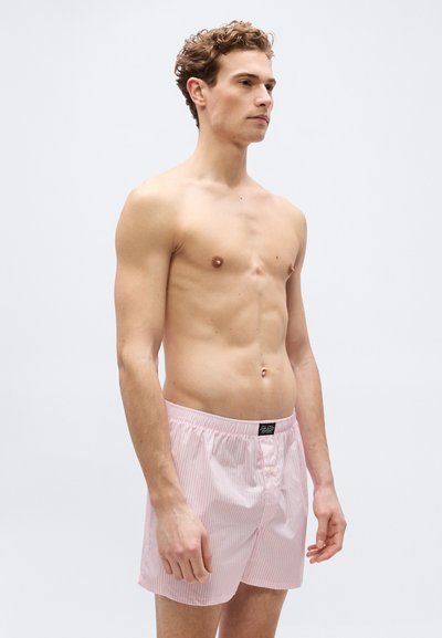 Άνδρας χωρίς πουκάμισο με σγουρά μαλλιά, ντυμένος με ροζ και άσπρα ριγέ σορτς μποξ, στέκεται μπροστά σε ένα απλό φωτεινό φόντο, κοιτάζοντας προς τα δεξιά.