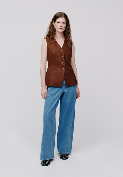 Gilet in lino marrone con cinque bottoni grandi, abbinato a jeans blu a gamba larga. Scarpe nere a pois completano l'outfit.