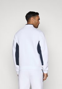 Fehér pulóver tengerészkék hálós oldalsávokkal, álló gallérral, bordás mandzsettával és szegéllyel. Simaság és sportos dizájn jellemzi.