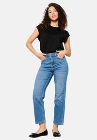 Top negro de manga corta, jeans de tiro alto en azul claro, de pierna recta, y bailarina negra, de pie con las manos en las caderas frente a un fondo blanco.