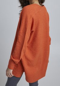 Oranžový převeliký pletený svetr s dlouhými rukávy, žebrovanými manžetami a rozparkem na spodním okraji. Měkká textura, volný střih a jemné žíhání po celé ploše.